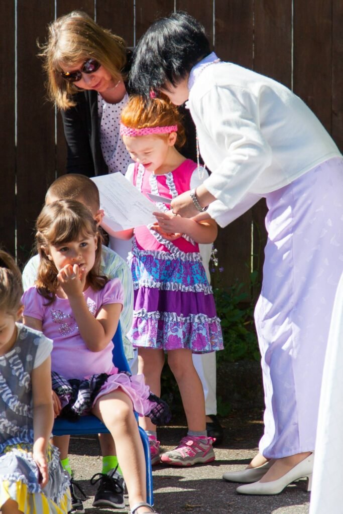 A small group of people at an outdoor event, with one person holding a young child. The setting includes a dark fence and bright sunlight.