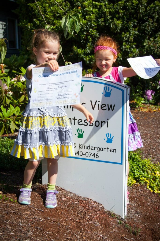 Two young children pose happily beside a Peace View Montessori Kindergarten sign, holding up papers in a sunlit yard.
