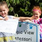 Two young children proudly displaying papers in front of an Olympic View Montessori sign in a garden setting.