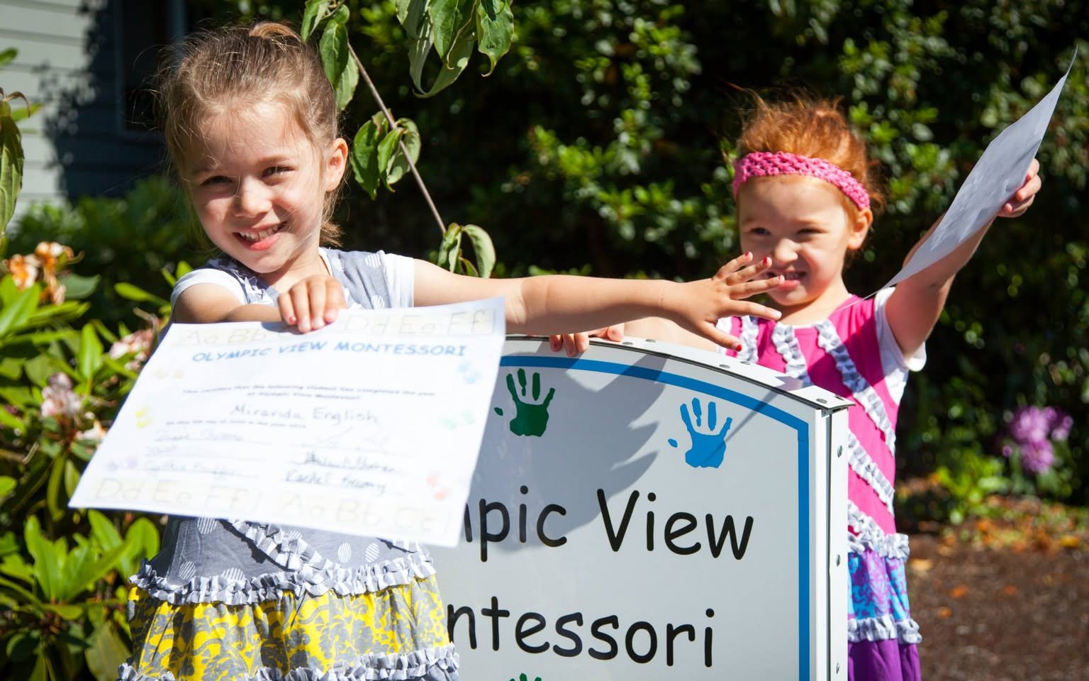 Two young children proudly displaying papers in front of an Olympic View Montessori sign in a garden setting.