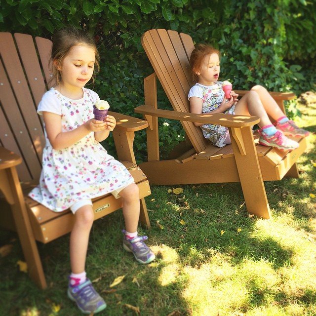 Two children sitting in wooden Adirondack chairs outside, each holding a small cup of ice cream. One child appears to be eating, while the other yawns.