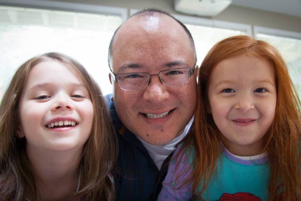 Three smiling people in a close-up selfie taken indoors. A man and two girls display happy expressions.