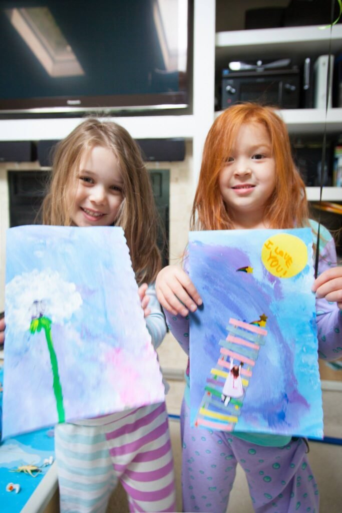 Two young children holding up colorful, handmade artwork featuring flowers and scenes with bright colors. Their artwork includes a dandelion and a figure on stairs with a sun.