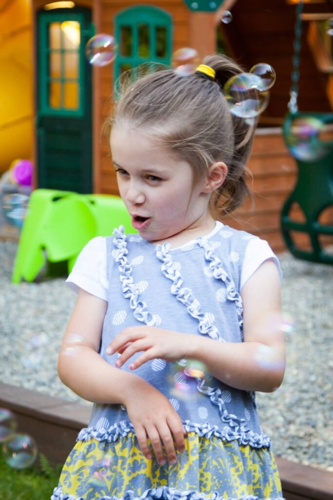 A child blows iridescent soap bubbles in a backyard featuring a playhouse and wooden swing, capturing a moment of childhood play.
