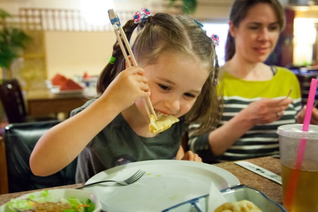 A young child uses chopsticks to eat food at a restaurant table with a family member nearby.