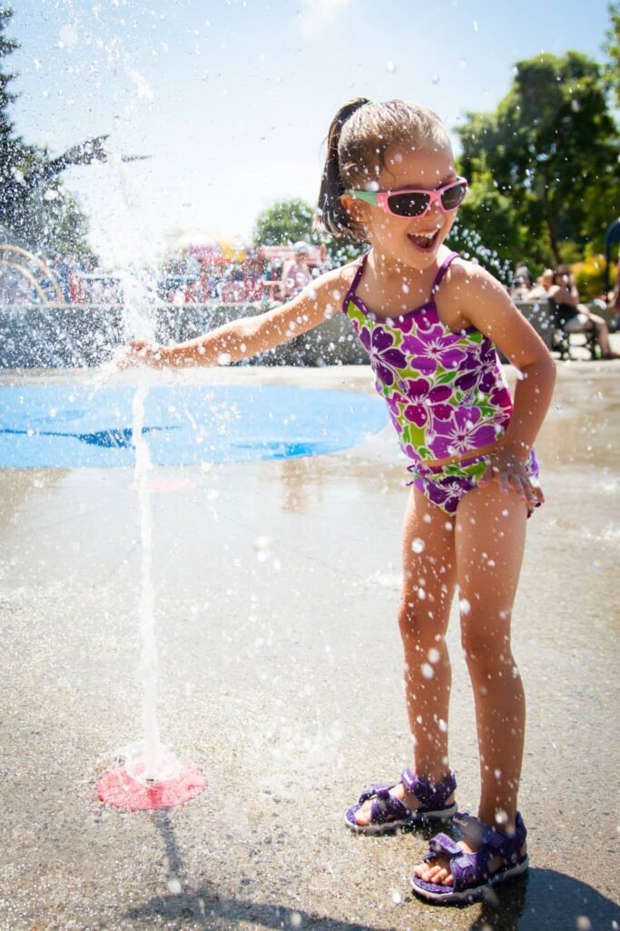 A child in a purple floral swimsuit stands by a water fountain, reaching out to touch the spray on a sunny day.