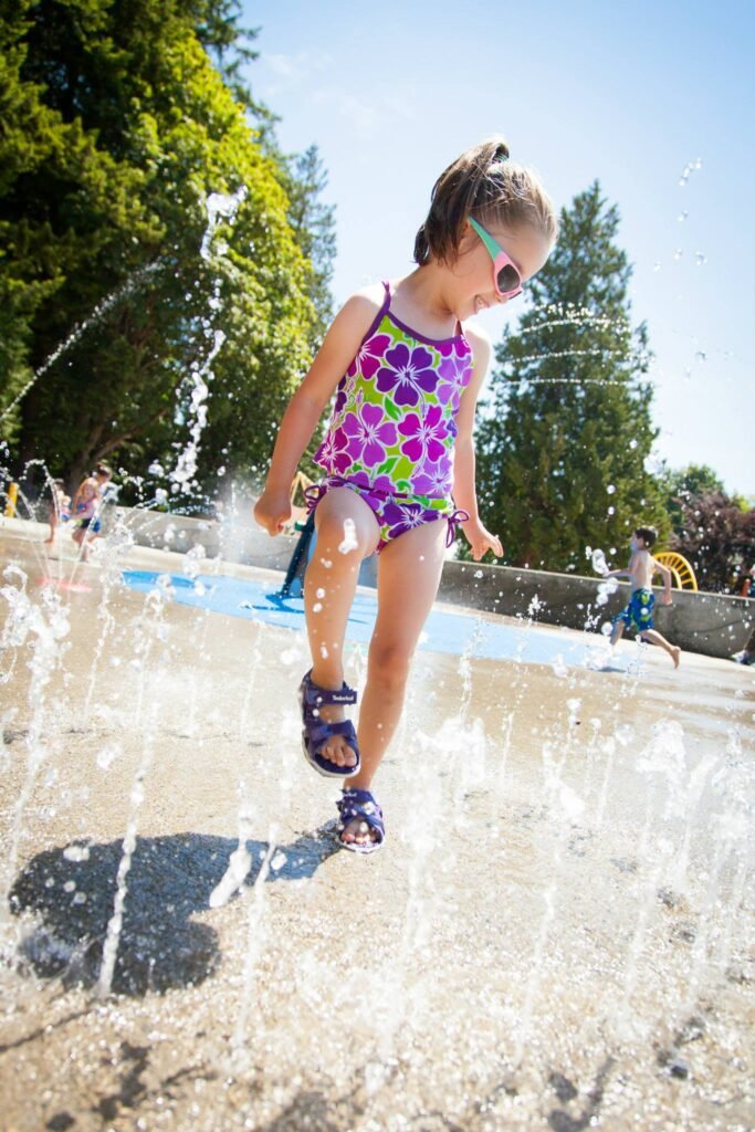 A child in a colorful swimsuit plays among the jets of an outdoor water fountain on a sunny day. The playful scene captures summer fun in a park setting.
