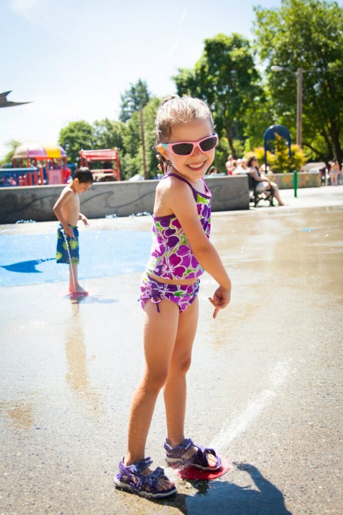 A child in a purple swimsuit enjoys playing with water sprinklers at a sunny outdoor water park. Other visitors can be seen enjoying the water features in the background.