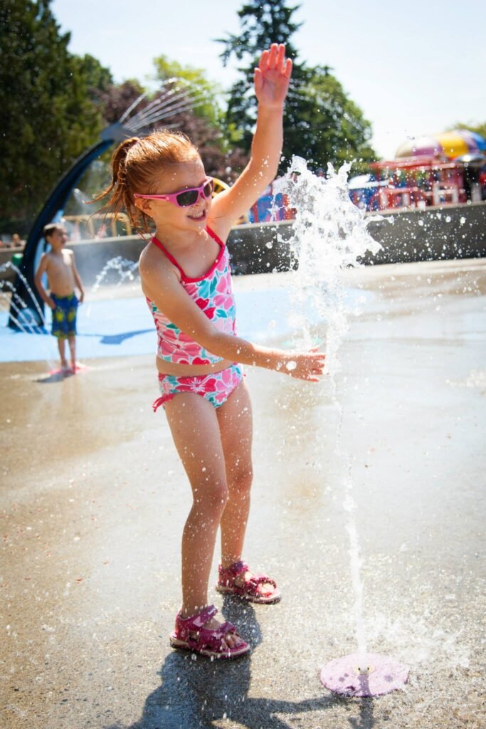 A child in a pink swimsuit splashes water at a splash pad with a colorful background.