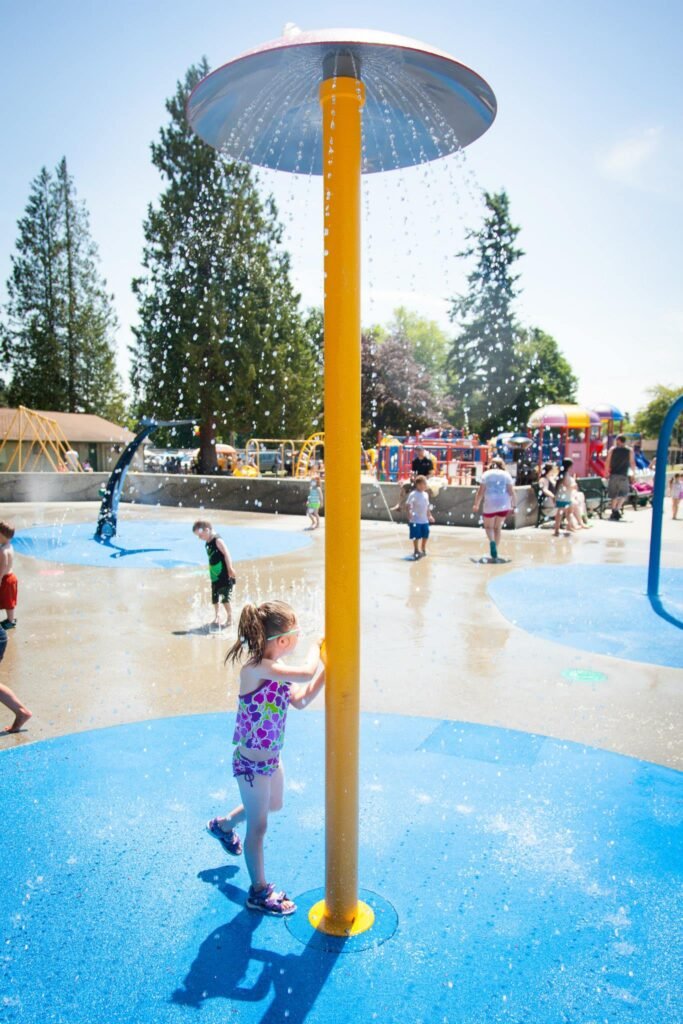 A yellow sprinkler tower sprays water in a splash park. Children play on the bright blue ground.