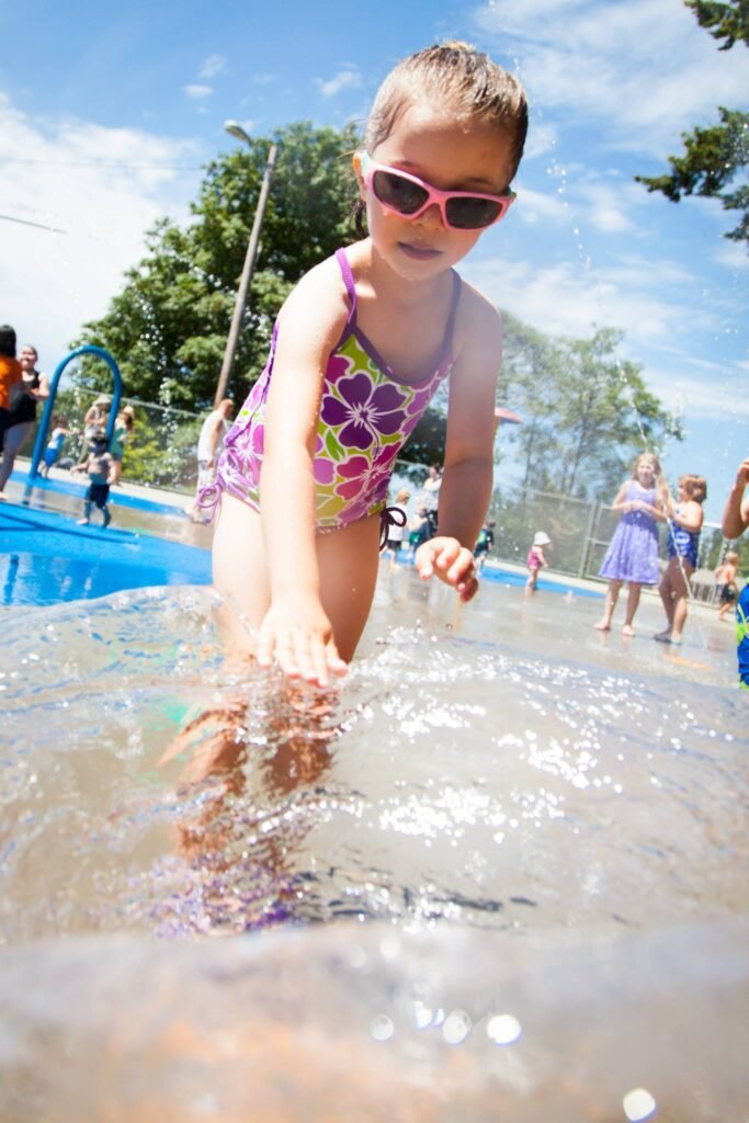 A child in a floral swimsuit plays in water at a sunny splash pad.