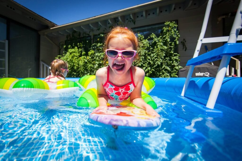 A child faces the camera while riding a small float in a backyard swimming pool. The setting includes a lifeguard stand, clear blue water, and bright daylight.
