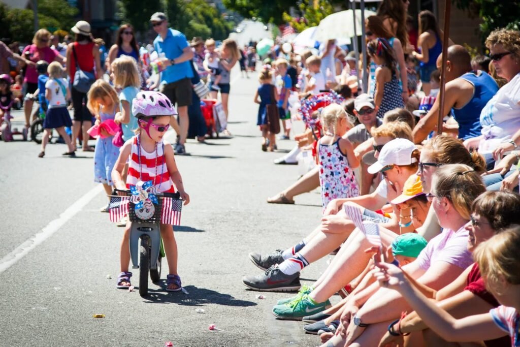 A young child in a helmet rides a decorated bicycle in a community parade, surrounded by an enthusiastic crowd.