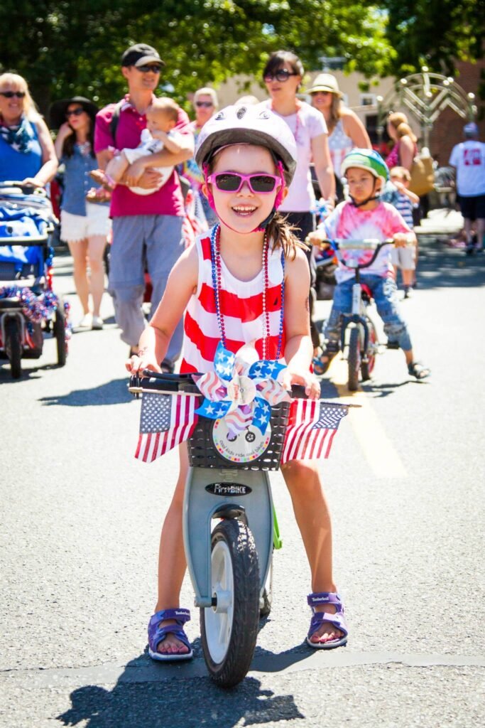 A young child rides a decorated bicycle with American flag accents during a sunny outdoor neighborhood bike event in summer.
