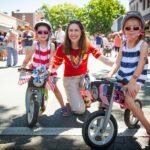 A smiling family posing with small bikes on a sunny street, with a crowd of people in the background enjoying a lively outdoor event.