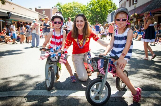 A smiling family posing with small bikes on a sunny street, with a crowd of people in the background enjoying a lively outdoor event.