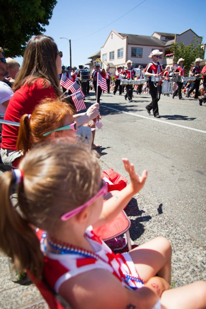 Children in patriotic attire watch a marching band during a sunny outdoor parade.