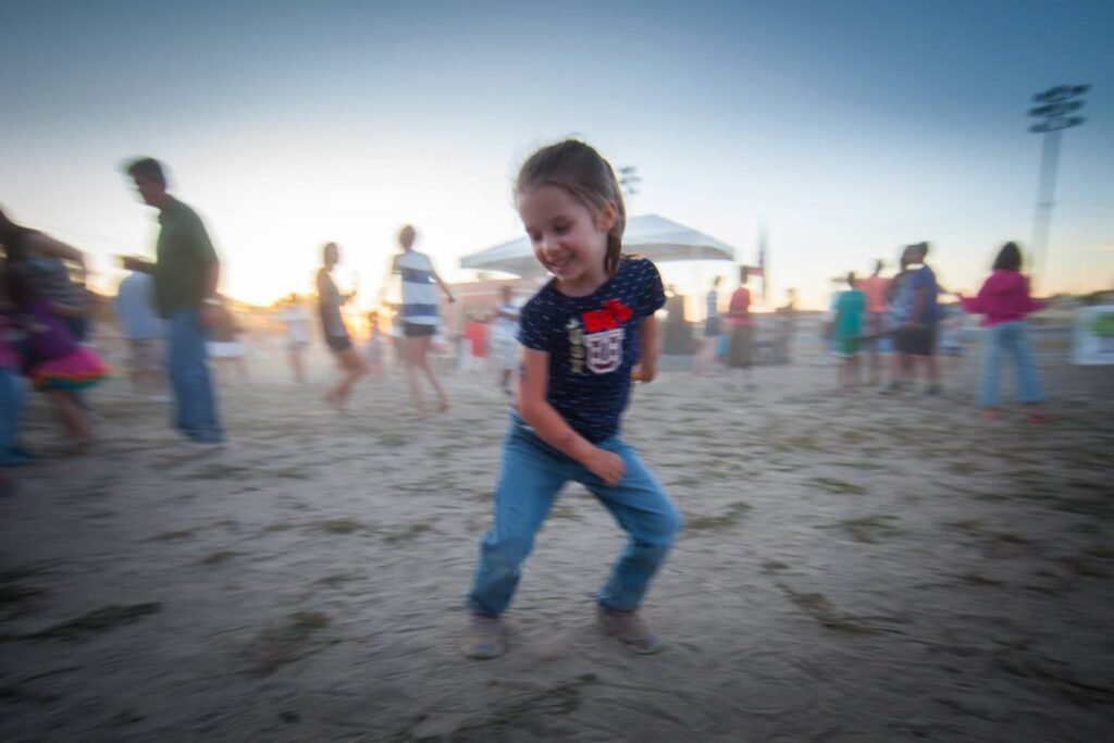 A child running in the soft sand on a crowded coastal beach with a sunset sky and tall umbrella in the background.