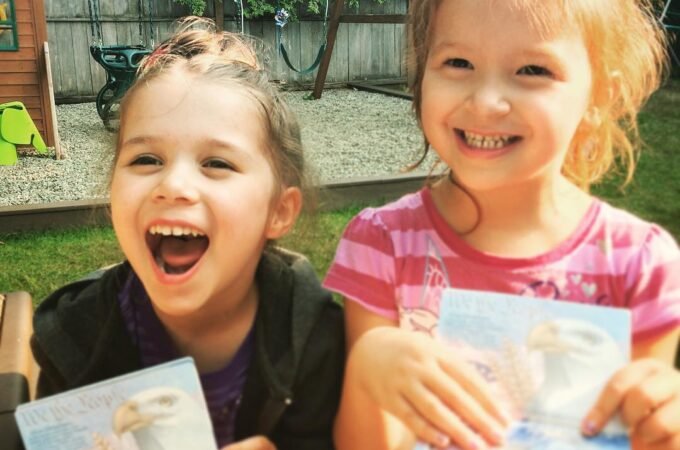Two young children smiling outdoors while holding homemade art cards. A close-up shows one child laughing joyfully.