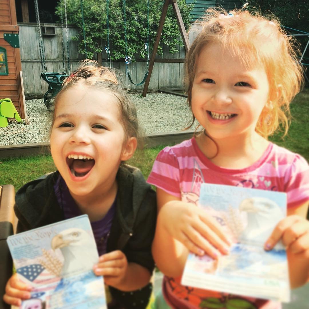 Two young children smiling outdoors while holding homemade art cards. A close-up shows one child laughing joyfully.