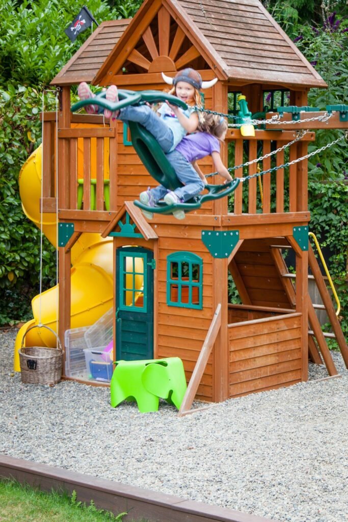 Children playing on a rope bridge attached to a wooden backyard playhouse with a slide.
