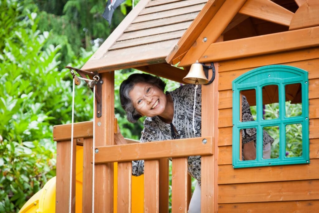 A smiling child hides and peeks out from the corner of a wooden playhouse with a teal window.