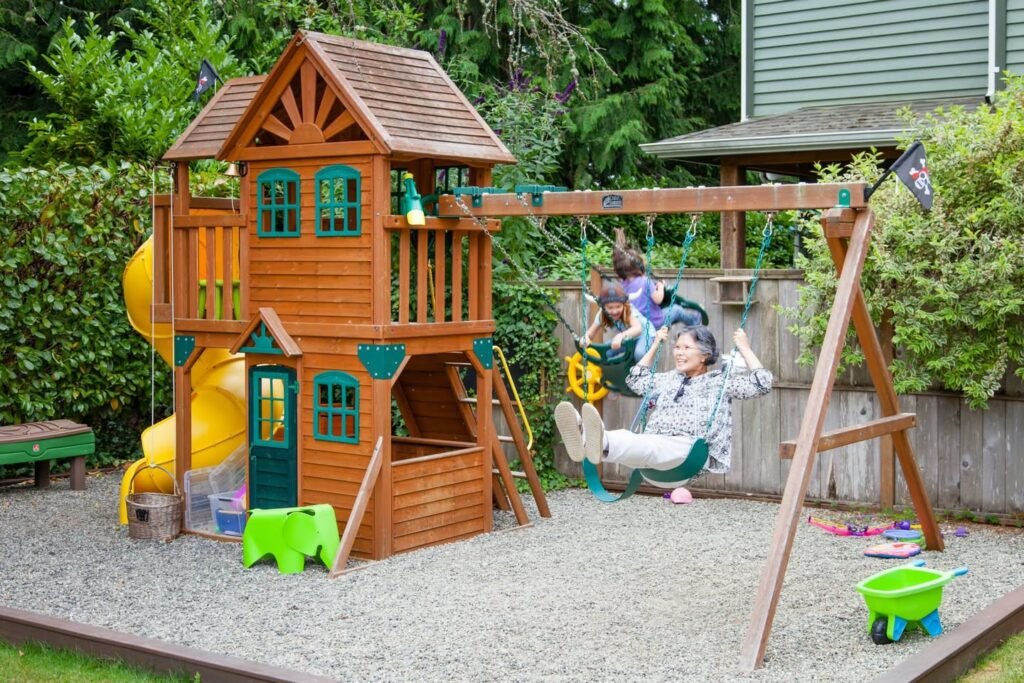 A large wooden playhouse with a yellow slide and swing set in a backyard. Gravel surface provides safe play area.