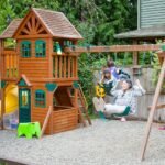 A large wooden playhouse with a yellow slide and swing set in a backyard. Gravel surface provides safe play area.
