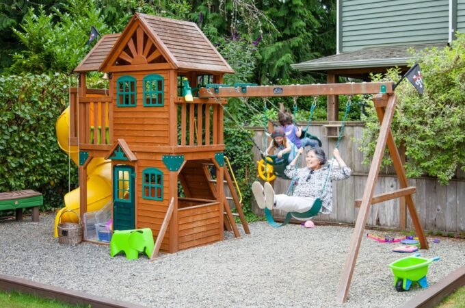 A large wooden playhouse with a yellow slide and swing set in a backyard. Gravel surface provides safe play area.
