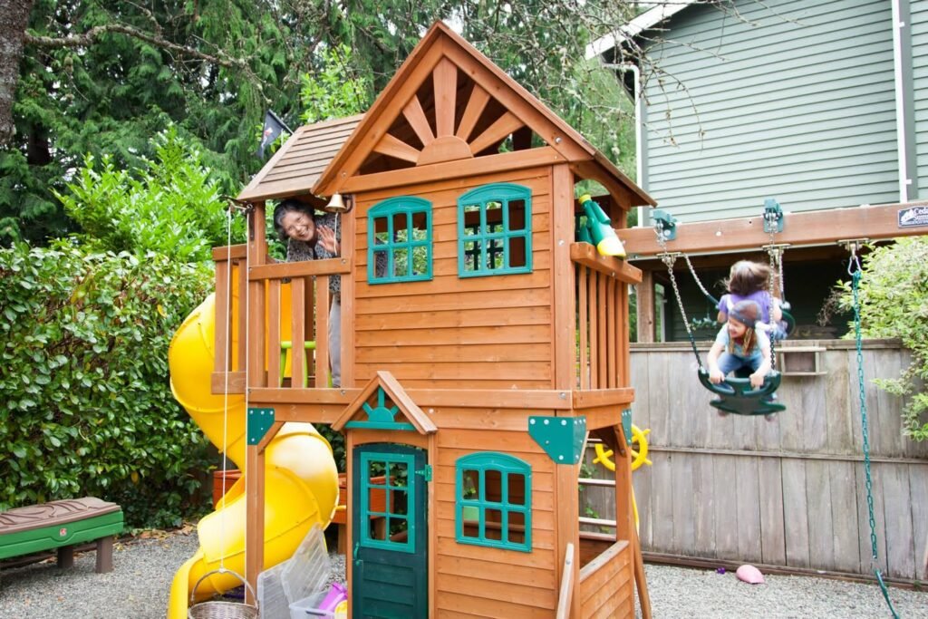 A wooden playhouse with a yellow slide and small swings in a backyard, set against greenery. Children enjoy the play area.