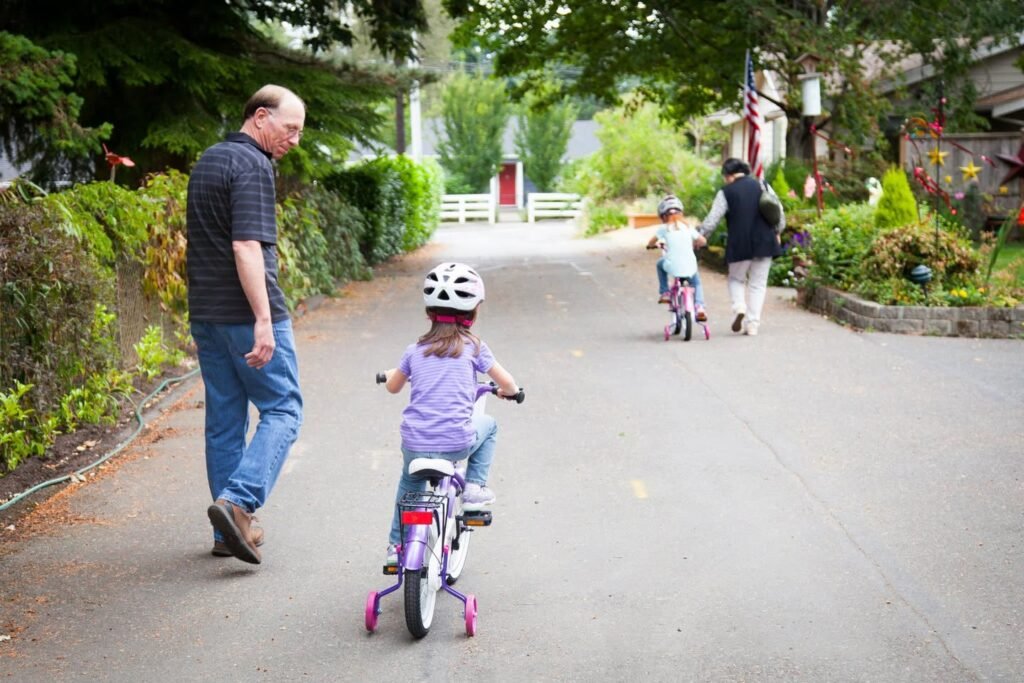 A child rides a balance bike with training wheels while an adult walks alongside. A second child practices further down the paved road.