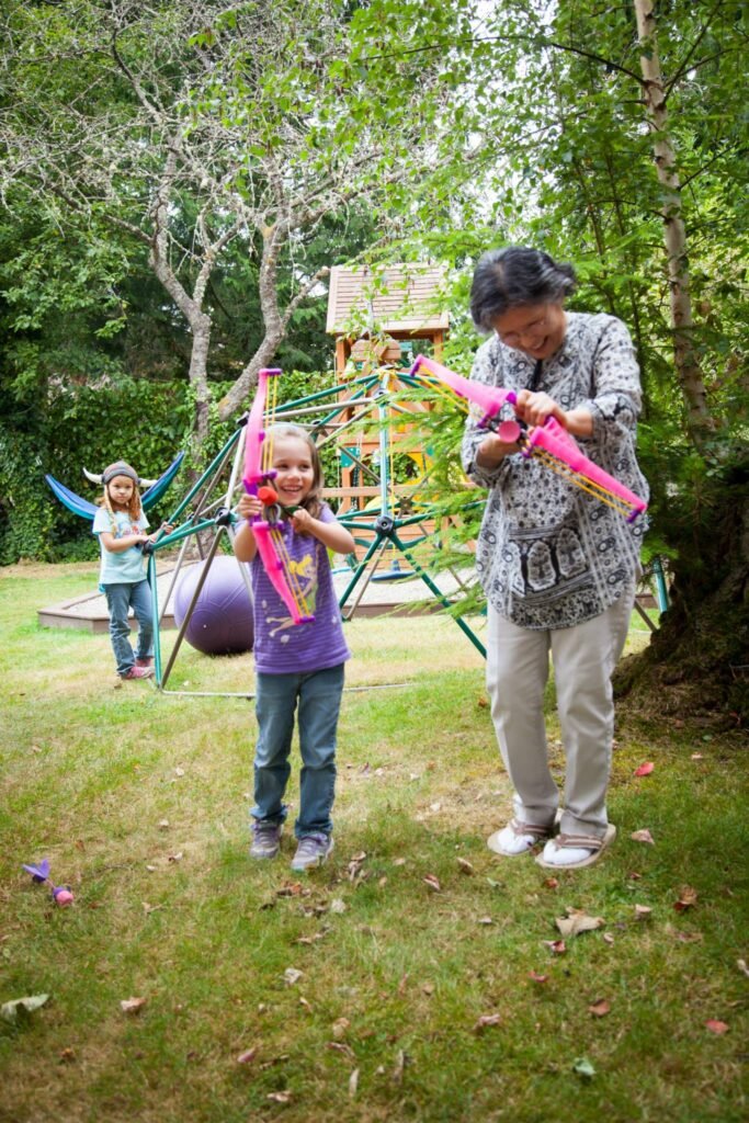 An adult and two children play with pink toy bows in a backyard. Nearby there is a tire swing and lush green trees.