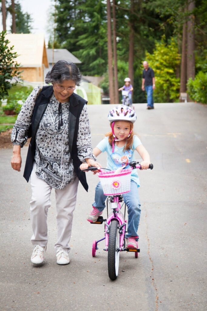 A child on a training bike with an adult nearby, surrounded by trees.