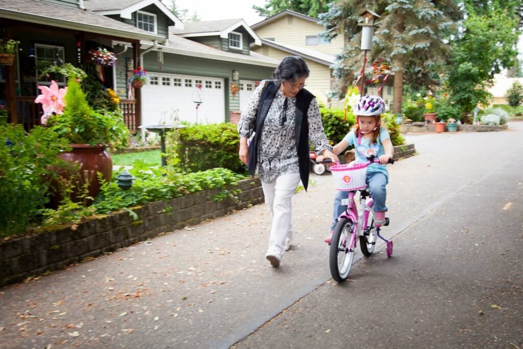 A child with a helmet riding a bicycle with training wheels while an adult walks alongside for support.