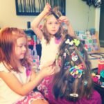 Two young children styling someone's long brown hair filled with colorful decorative bows. A bathroom setting with hair accessories arranged in the background.
