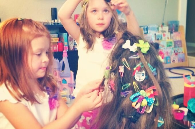 Two young children styling someone's long brown hair filled with colorful decorative bows. A bathroom setting with hair accessories arranged in the background.