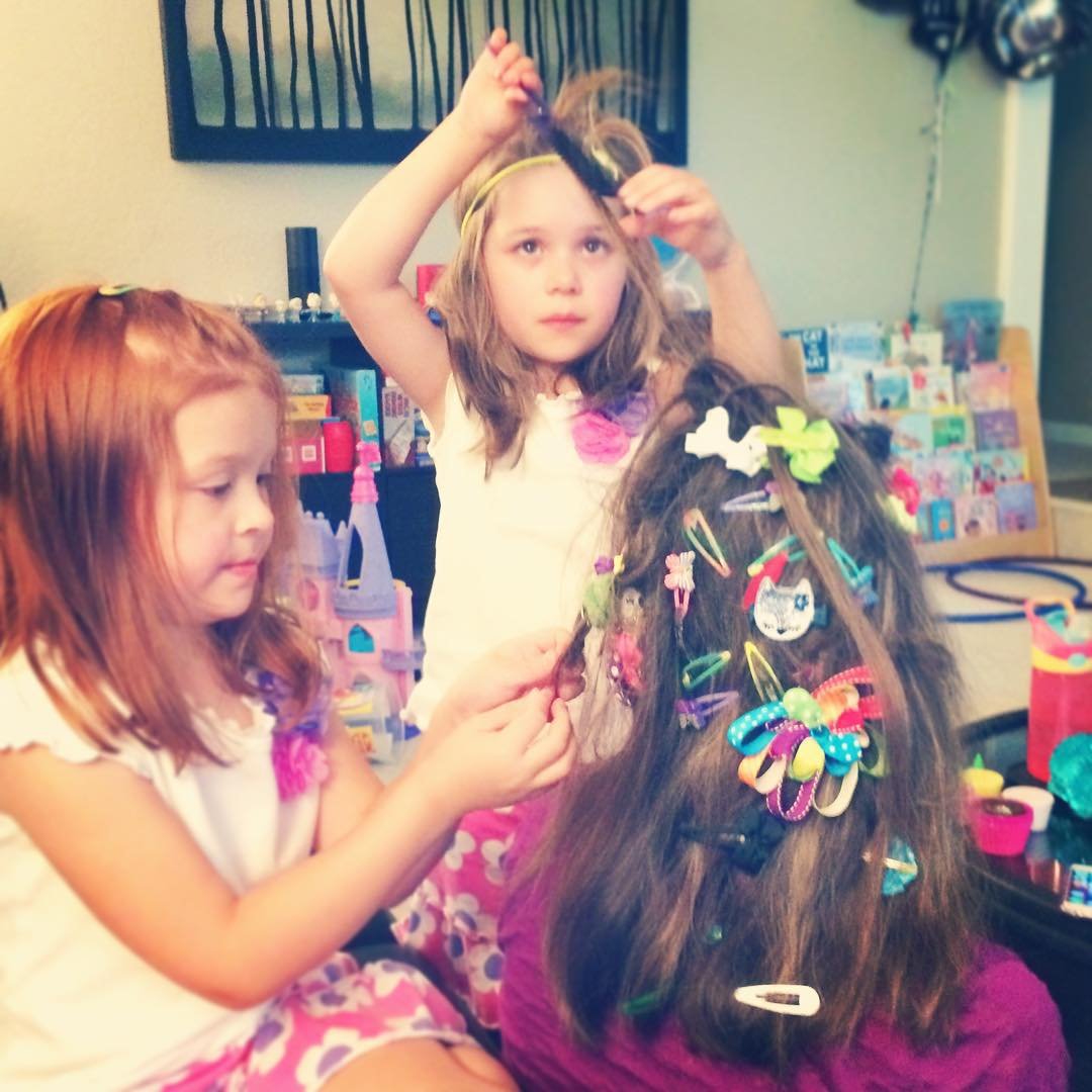 Two young children styling someone's long brown hair filled with colorful decorative bows. A bathroom setting with hair accessories arranged in the background.