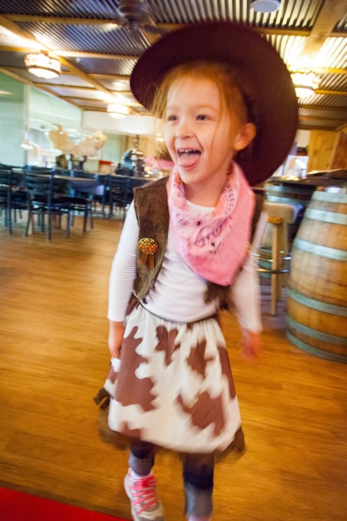 A child wearing a cowboy hat and cow-print skirt smiles near wooden wine barrels in a rustic tasting room.
