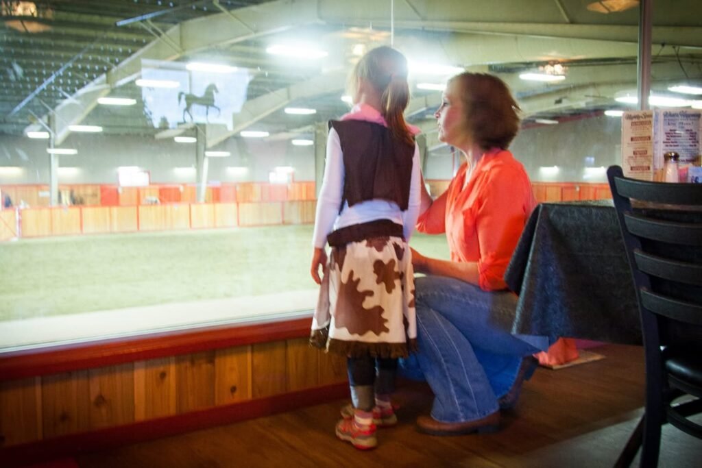 A small child and an adult look out at a horse in an indoor riding arena. They are seated near a large window overlooking the track.