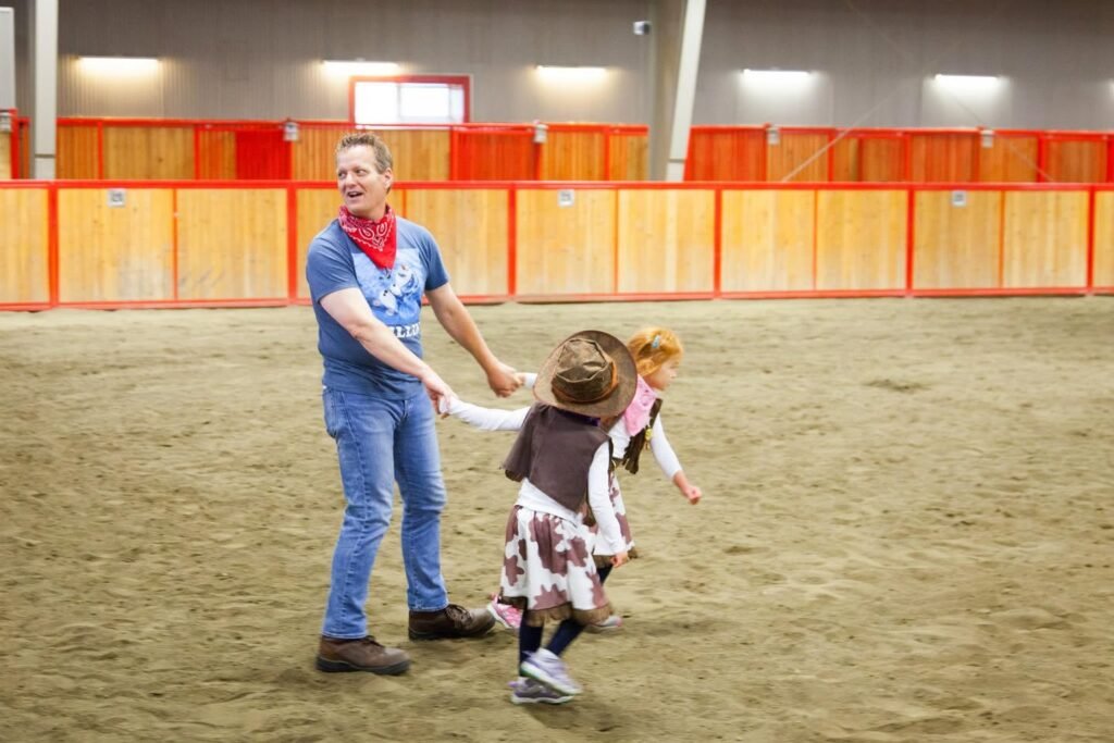 A joyful family dance session inside an indoor riding arena with wooden fencing and sandy floors.