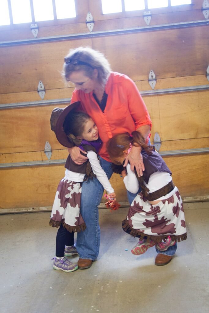 A woman helps two young kids dressed in cow costumes pose indoors. The setting features rustic wooden barn walls.