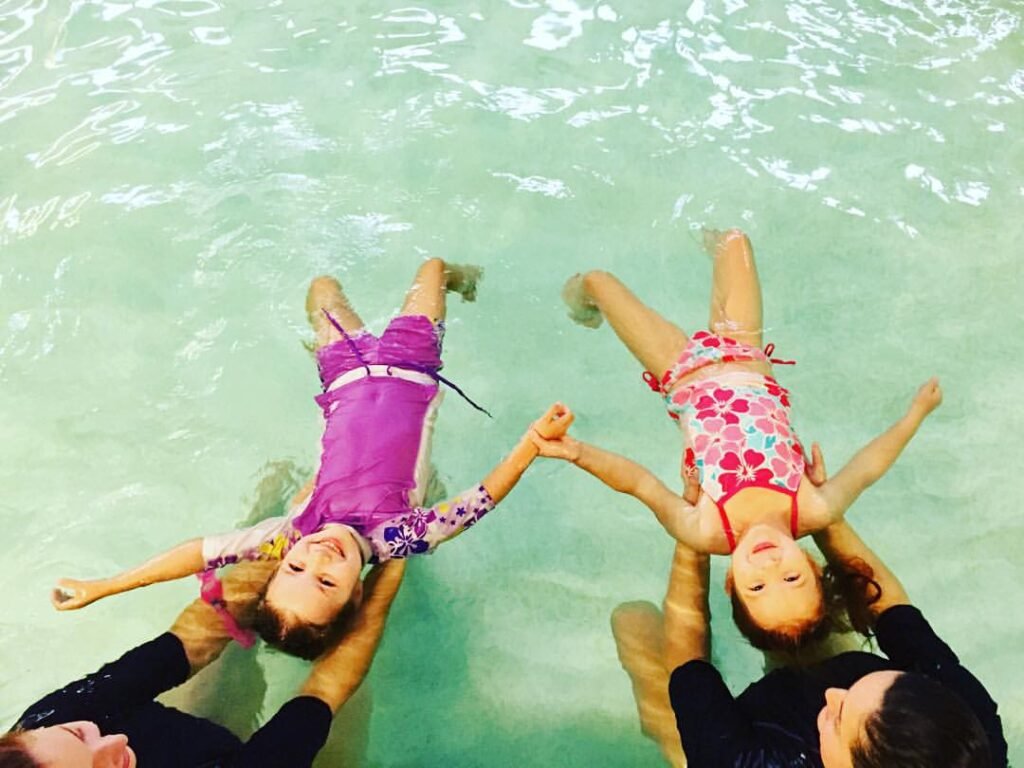 A family enjoys swimming together in a clear pool with turquoise water. The children are being held by adults while holding hands above the surface.