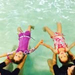 A family enjoys swimming together in a clear pool with turquoise water. The children are being held by adults while holding hands above the surface.