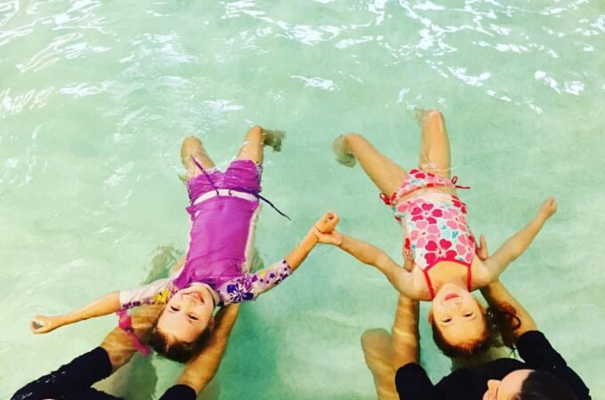 A family enjoys swimming together in a clear pool with turquoise water. The children are being held by adults while holding hands above the surface.