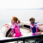 Two children wearing life jackets sit at the bow of a boat gliding over calm water with distant hills in the background.