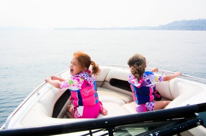 Two children wearing life jackets sit at the bow of a boat gliding over calm water with distant hills in the background.
