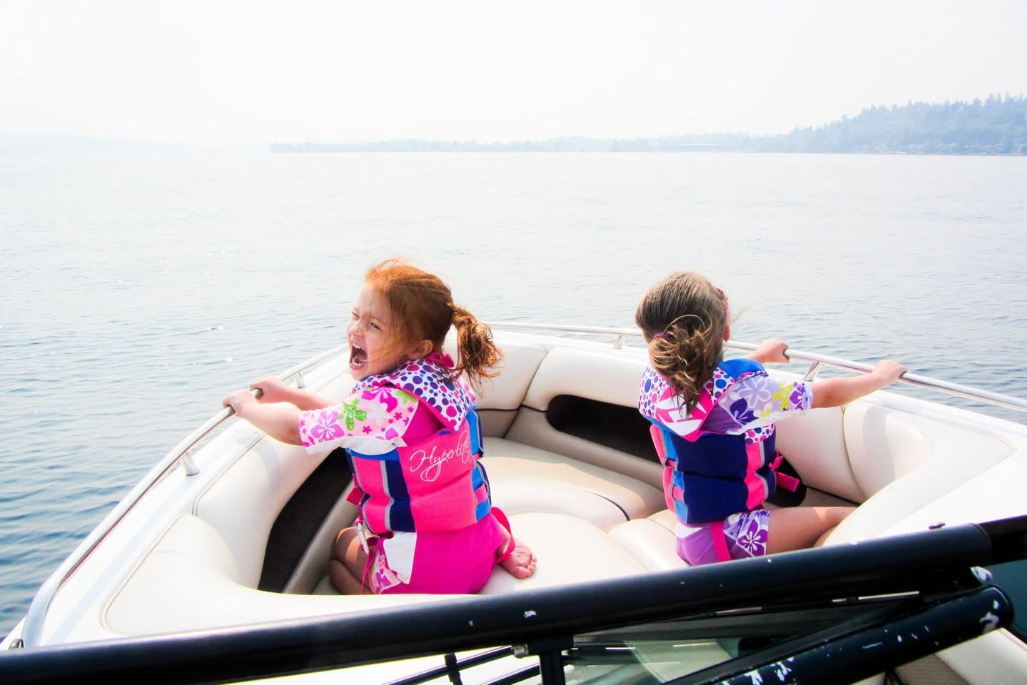 Seattle Boating Two children wearing life jackets sit at the bow of a boat gliding over calm water with distant hills in the background.