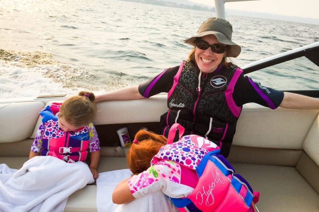 A smiling woman in a sun hat and life jacket sits on a boat with two children also wearing brightly colored life jackets. The calm water in the background suggests a peaceful day out on the lake.