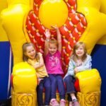 Three girls sitting and posing joyfully on a bright yellow and red inflatable throne against a colorful bouncy castle.