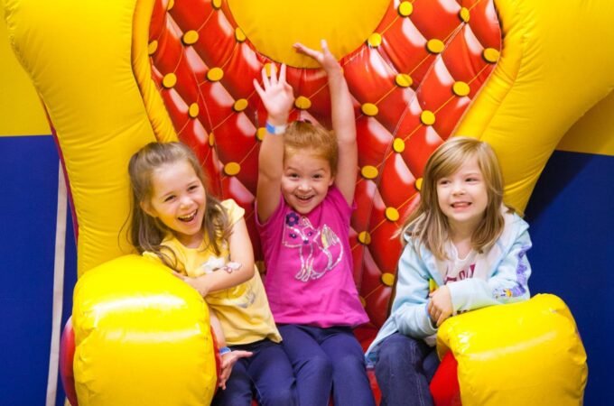 Three girls sitting and posing joyfully on a bright yellow and red inflatable throne against a colorful bouncy castle.